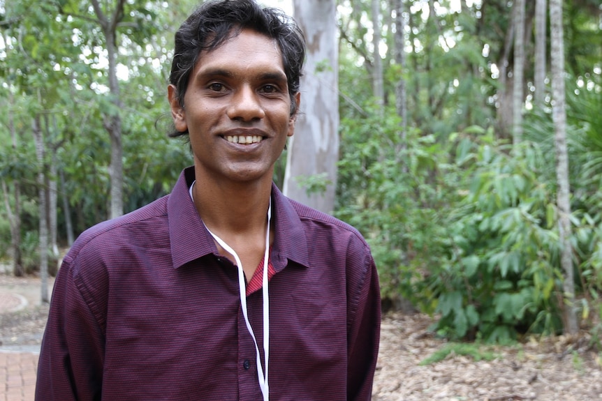 A young man stands on a footpath and smiles at the camera.
