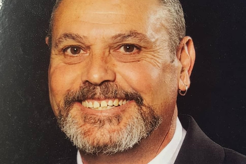 A professional headshot of a man with a beard, wearing a black suit smiling in front of a black backdrop