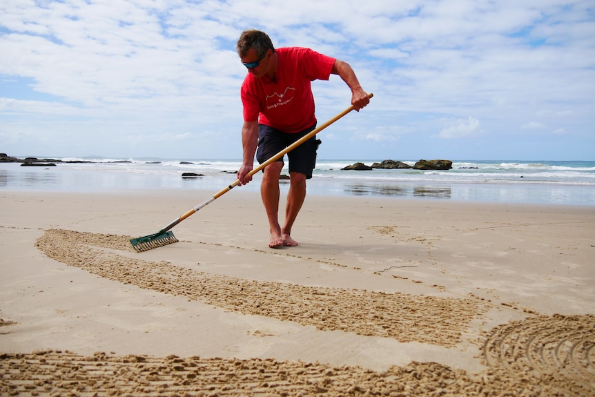 A man uses a rake to create patterns on the sand at a beach, a close up of the lines in the sand.