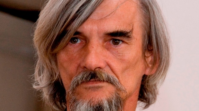 A man dressed in black with long hair and a beard looks past the camera.