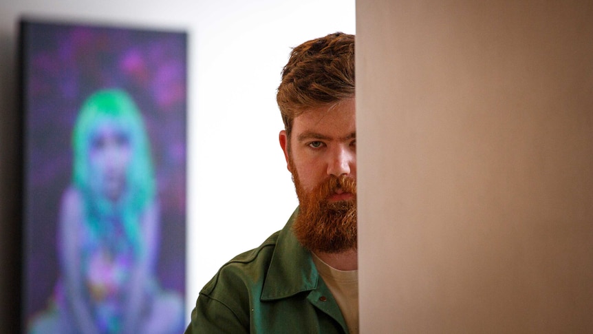 A young bearded man stares out from behind a wall. In the background is a bright purple and green artwork of a person.