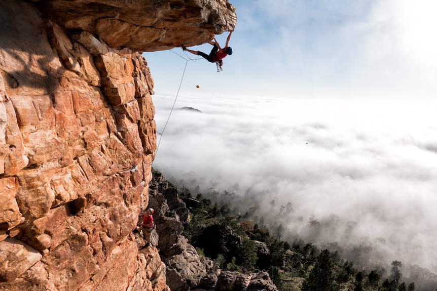 a woman climbs around a really high overhang, as a man belays her below