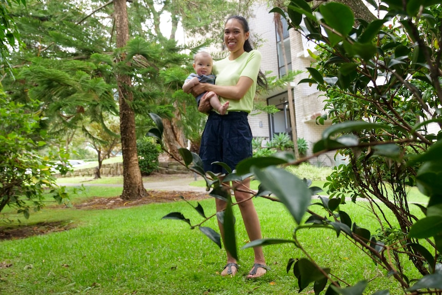 A woman in a green t-shirt holds a baby, standing on grass and surrounded by trees.