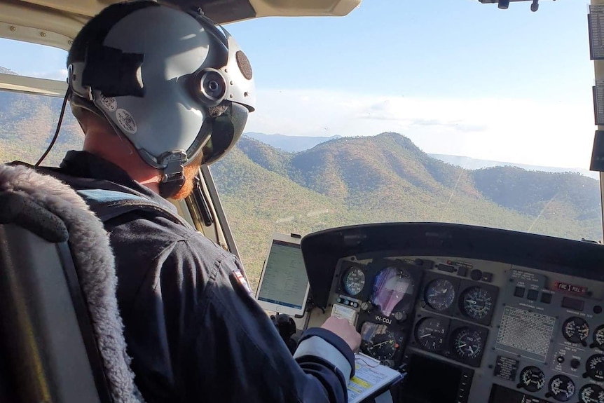 View from inside a helicopter, over the shoulder of a pilot wearing a helmet and headset.