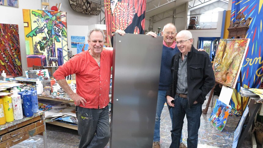 Three men in an art studio holding up a stainless steel fridge door