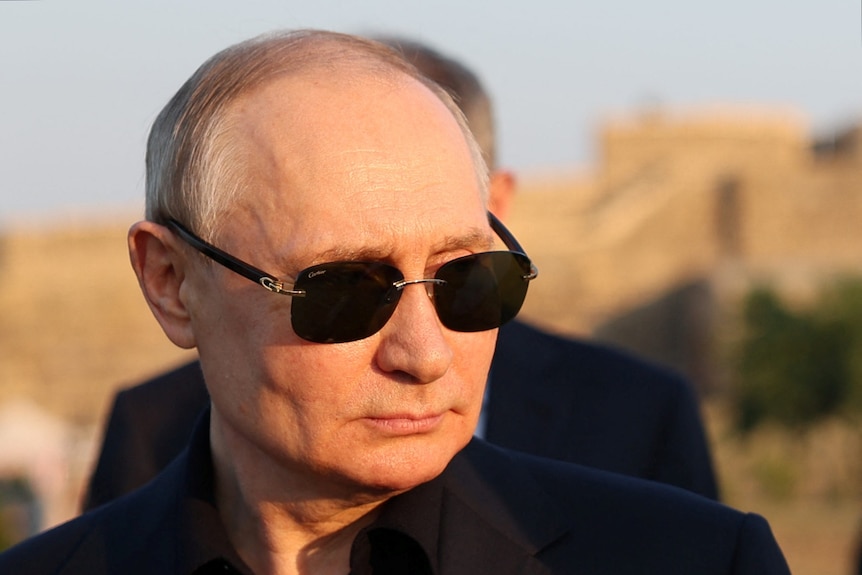 Le visage de Vladimir Poutine est éclairé par la chaleur du soleil.  Il porte des lunettes de soleil foncées et une veste noire sur une chemise noire