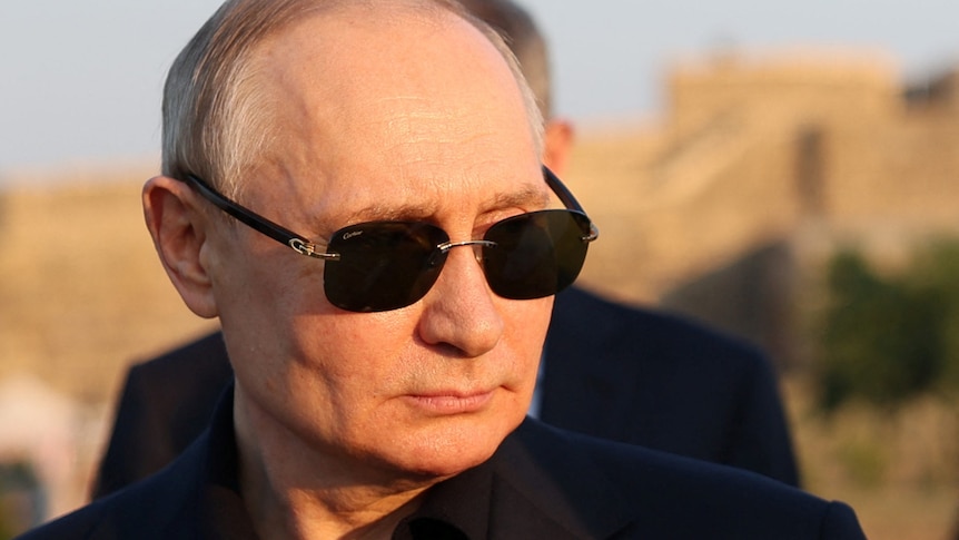 Vladimir Putin's face is lit by warm sunlight. He wears dark sunglasses and a black jacket on black shirt