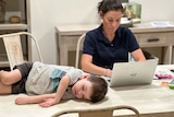 Toddler in T-shirt, shorts asleep on a dining table next to a woman wearing a blue tee with company labels, works on a computer.