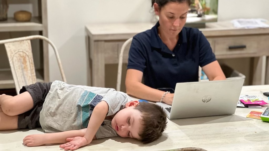 Toddler in T-shirt, shorts asleep on a dining table next to a woman wearing a blue tee with company labels, works on a computer.