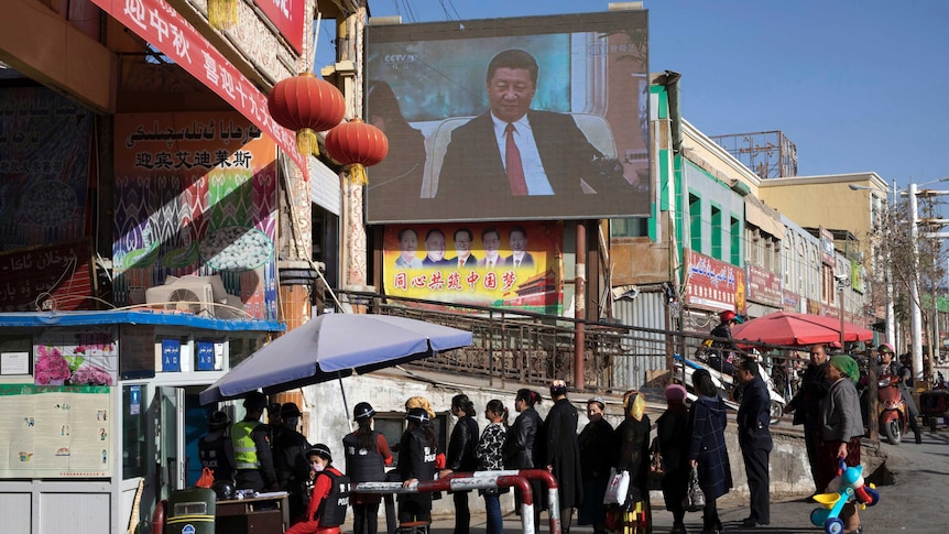 People walk through a security checkpoint into the Hotan Bazaar where a screen shows Chinese President Xi Jinping.