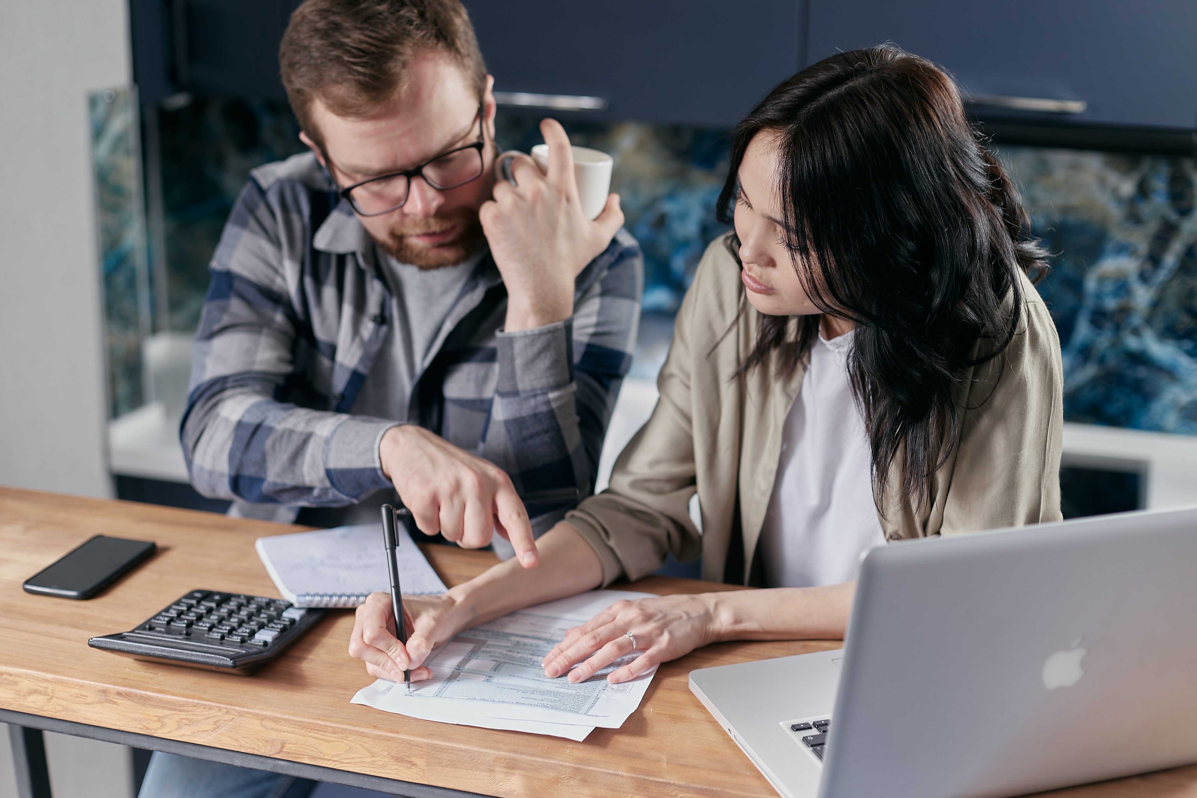 A man and woman sit a bench, looking at paper bills, with a calculator and laptop next to them
