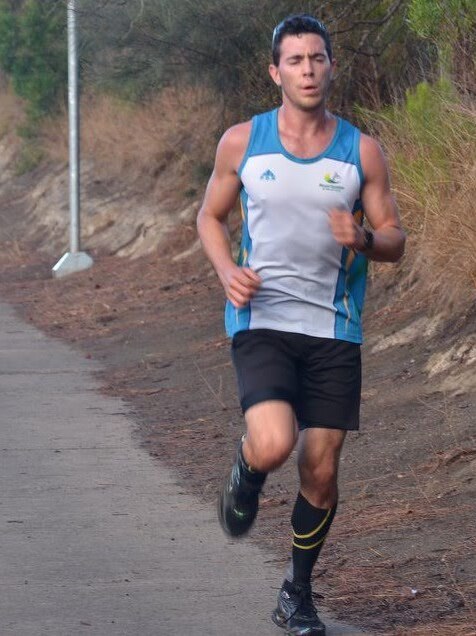 A young, fit man jogs down a footpath. He wears a blue and white singlet and black shorts and his eyes closed.