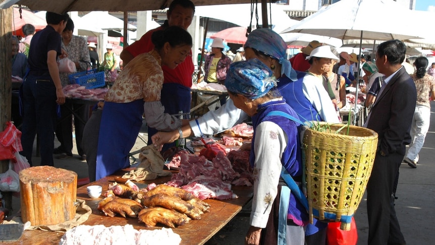 Chinese women buy and sell meat over a table laden with different cuts of pork. One lady has a basket on her back.