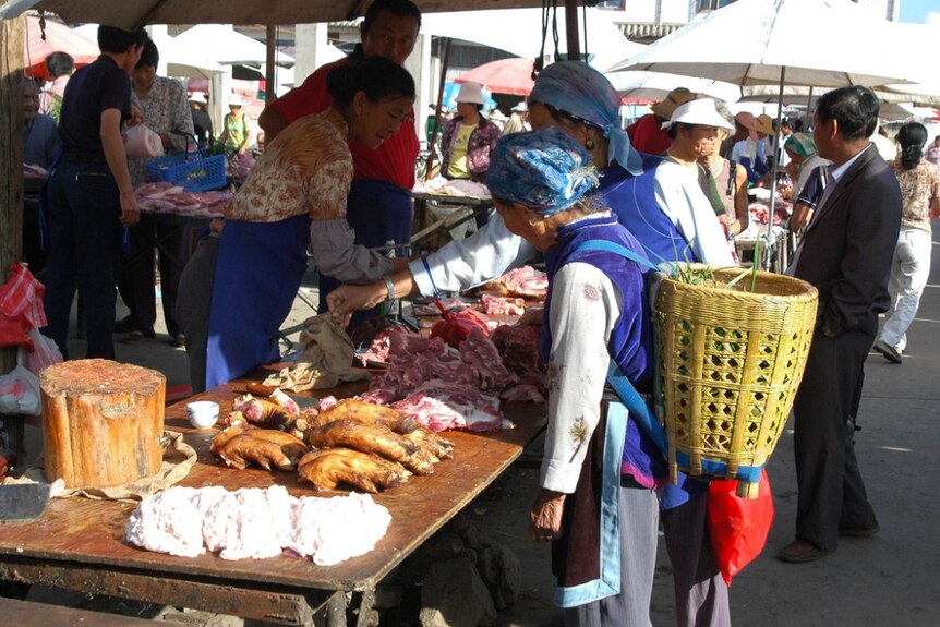 Chinese women buy and sell meat over a table laden with different cuts of pork. One lady has a basket on her back.