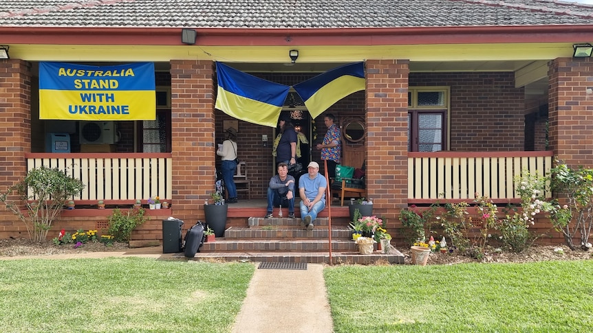 The front of a convent with people sitting on the steps and three Ukrainian flags hanging.