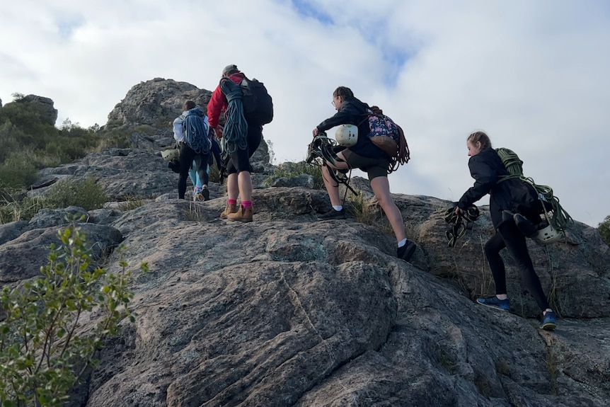 four girls carrying backpacks hike up rocky terrain.