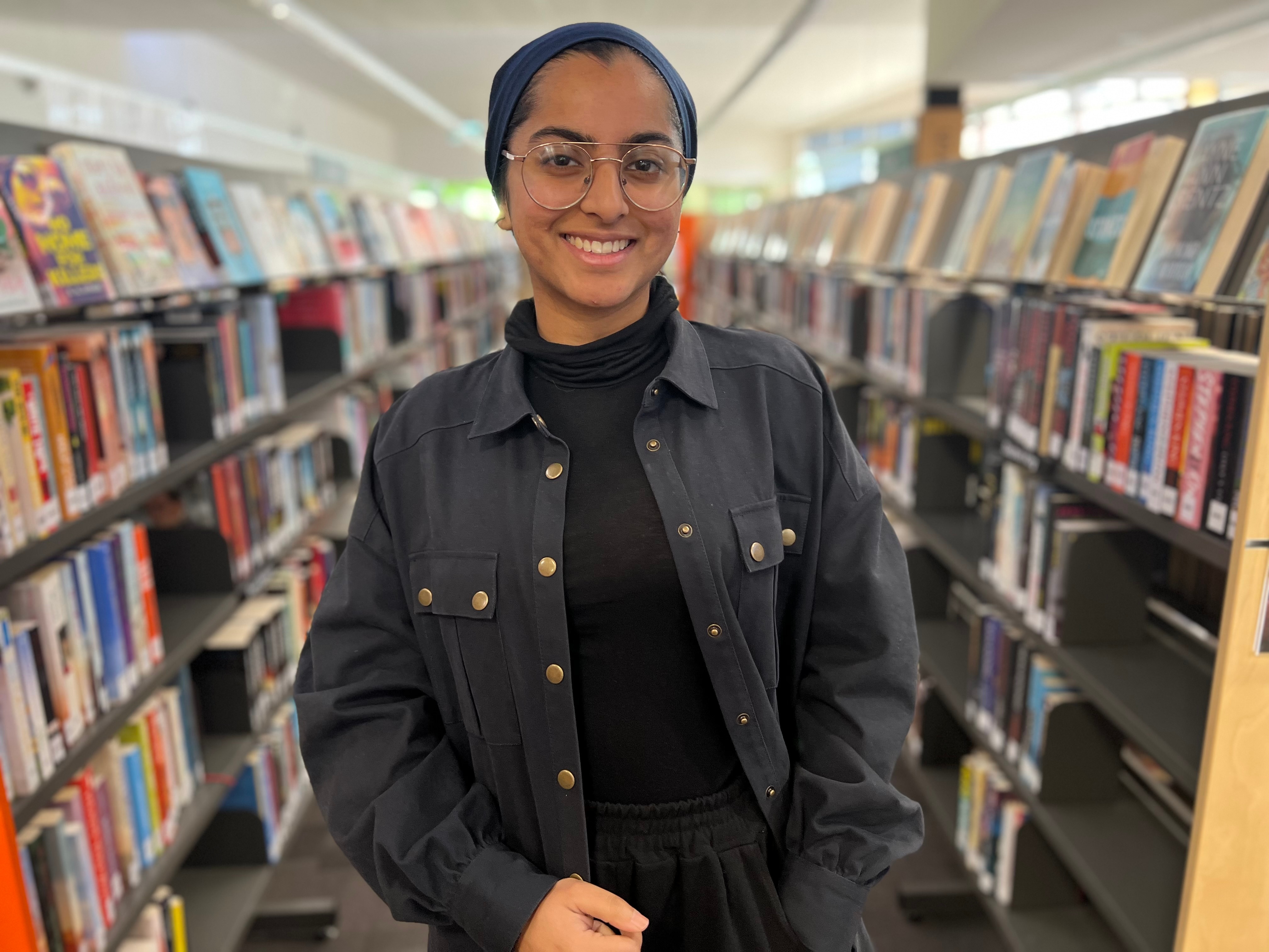A woman wearing a hijab standing near two bookshelves.