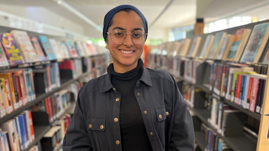 A woman wearing a hijab standing near two bookshelves.