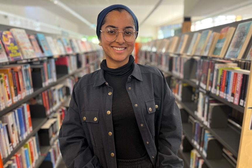 A woman wearing a hijab standing near two bookshelves.