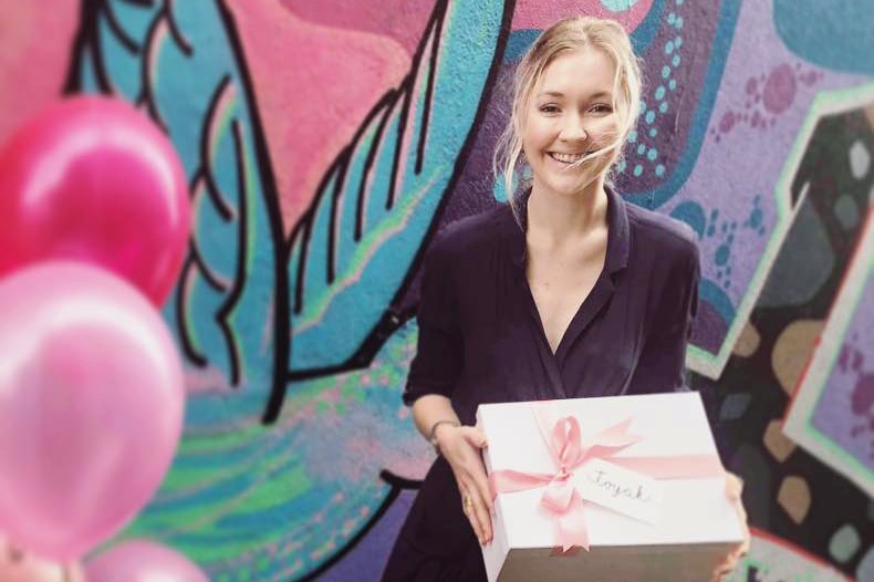 Toyah Cordingley smiles as she hold a wrapped present while standing in front of a mural.
