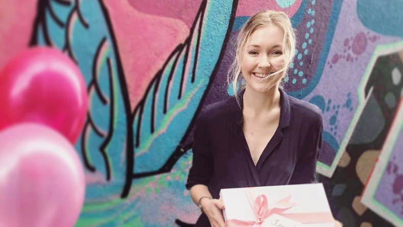Toyah Cordingley smiles as she hold a wrapped present while standing in front of a mural.