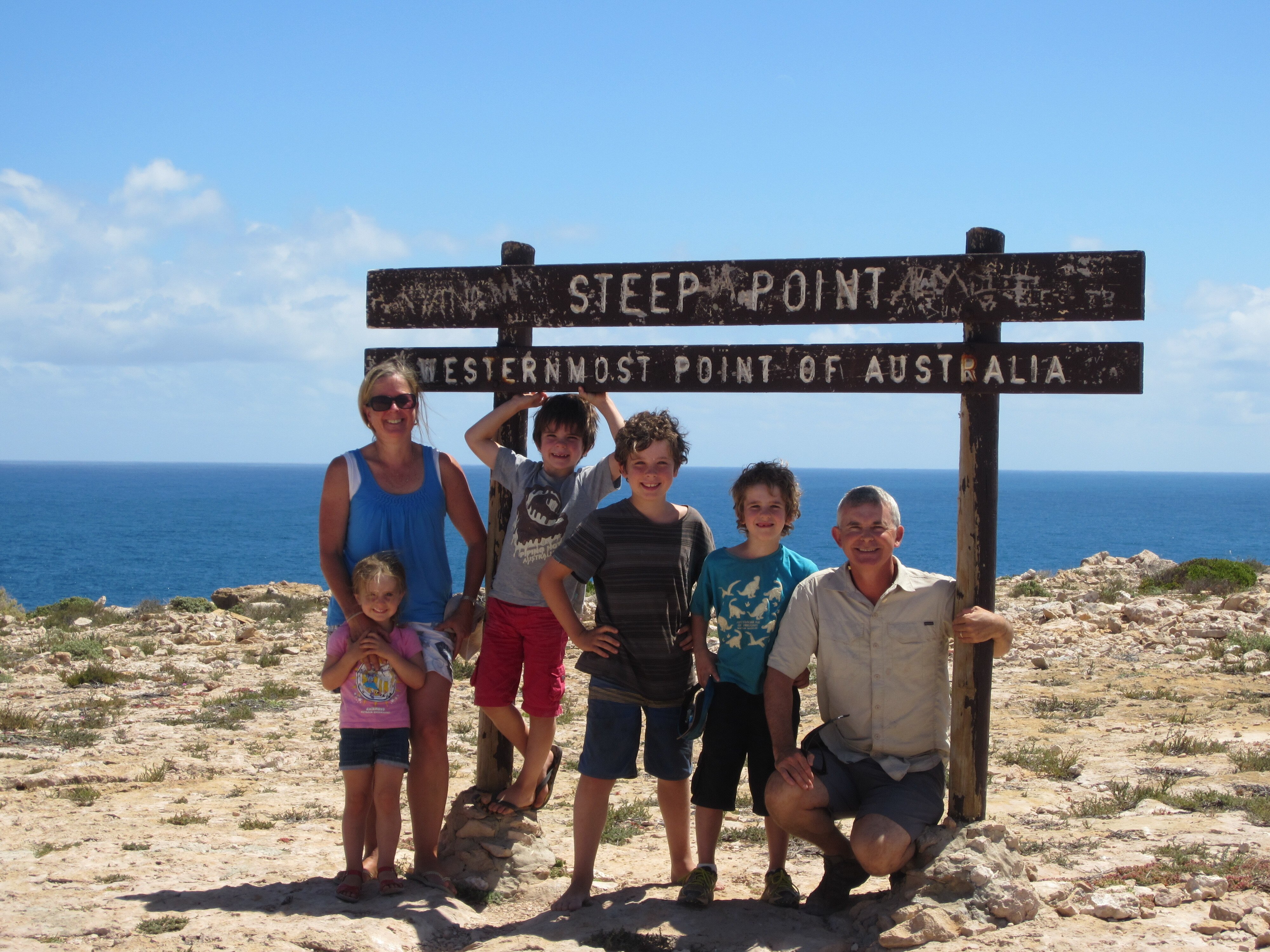 A family of 6 posed under a sign reading 'steep point' the 'westernmost point of Australia', with the ocean in the background.