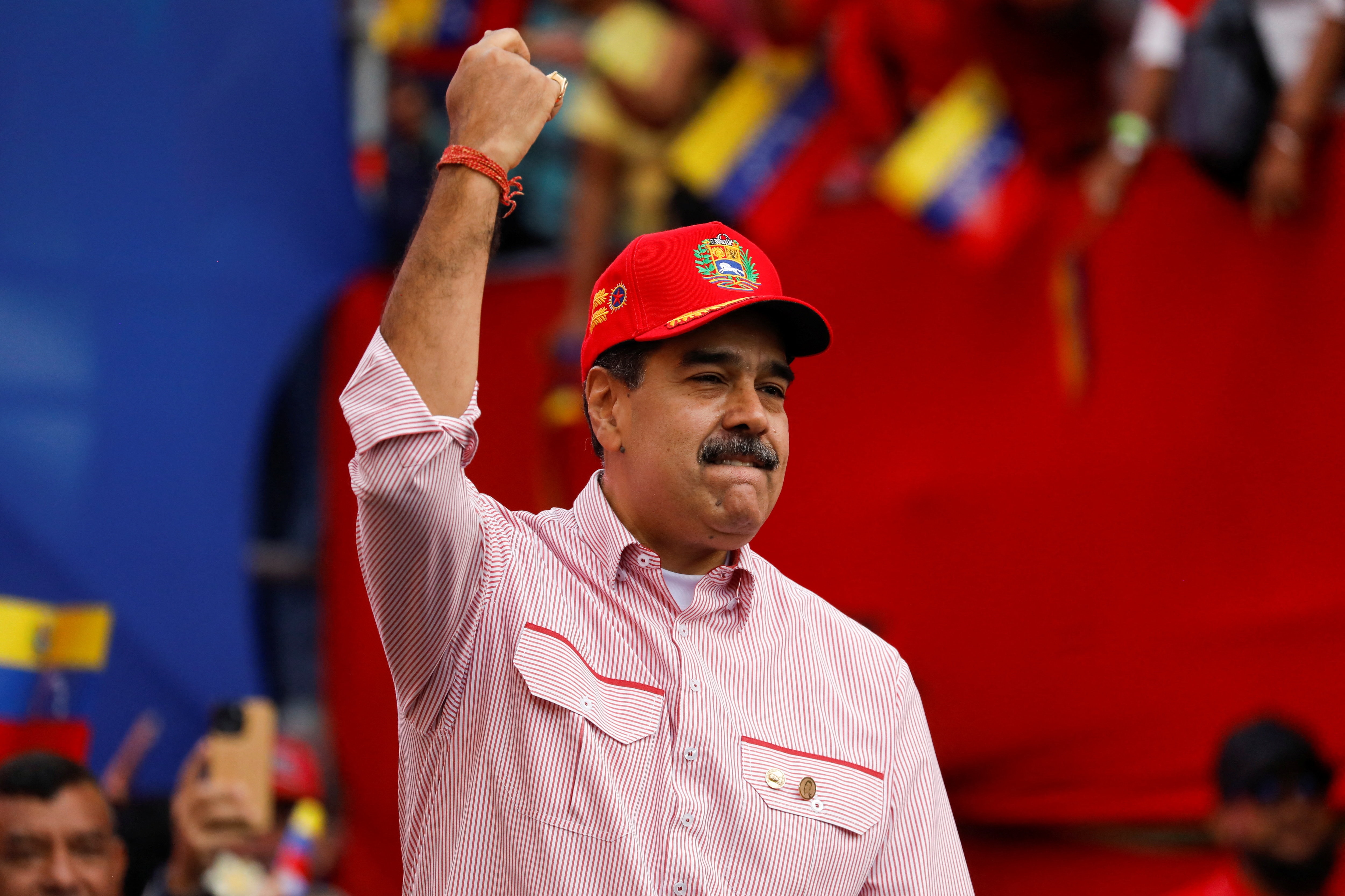 Nicolás Maduro sonríe y levanta el brazo derecho mientras viste una camisa a rayas rojas y blancas y una gorra roja.