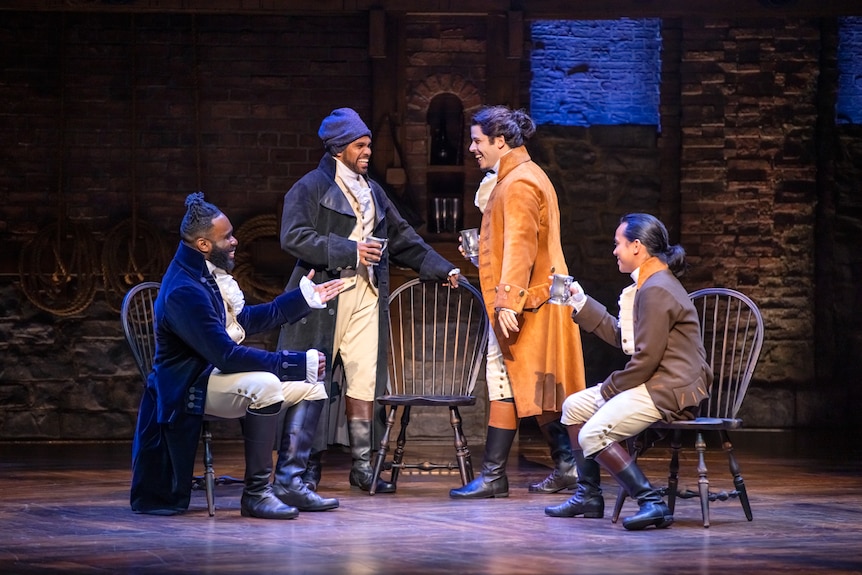 Four men in 18th century costume standing and sitting in group on stage, drinking and laughing.