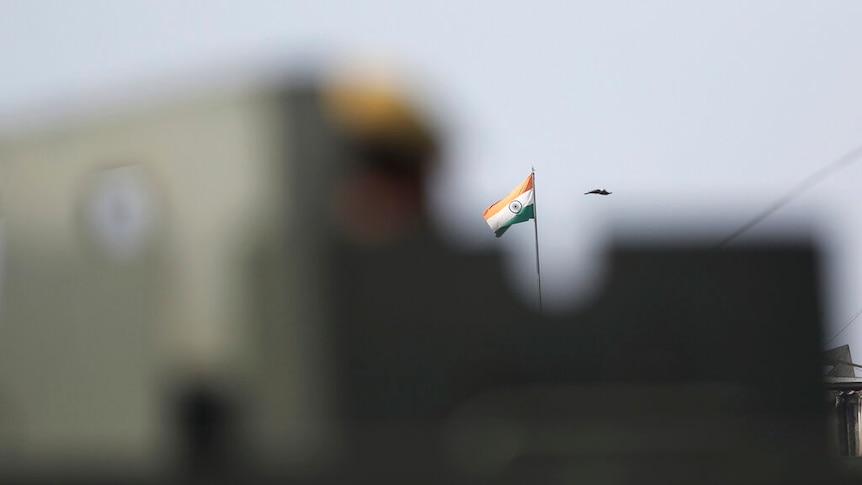 You view the Indian flag past a military vehicle which is out of focus.