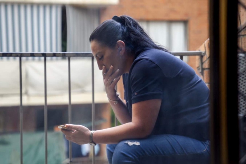 Woman with black hair sits outside on balcony smoking cigarette looking at phone with brick apartment building in background.