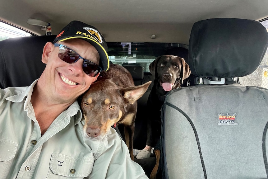 A  smiling man in a car with two dogs in the back seat. One dog puts its head on the man's shoulder. Man wears cap, green shirt.
