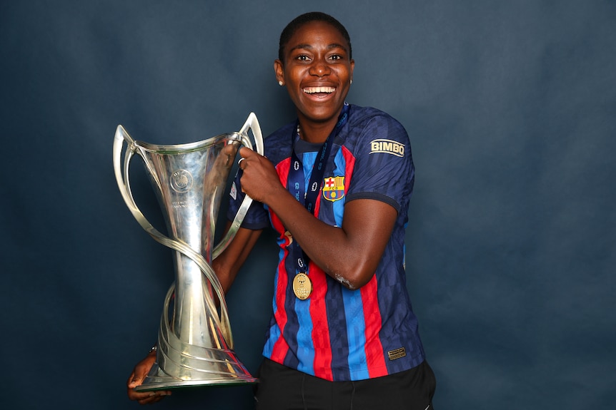 A soccer player wearing red and blue poses with a big silver trophy after winning a tournament