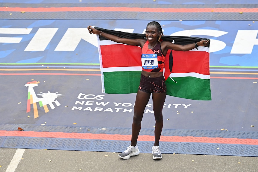 A female runner stands smiling on a roadway next to a sign saying 'New York City Marathon', holding a Kenyan flag behind her.