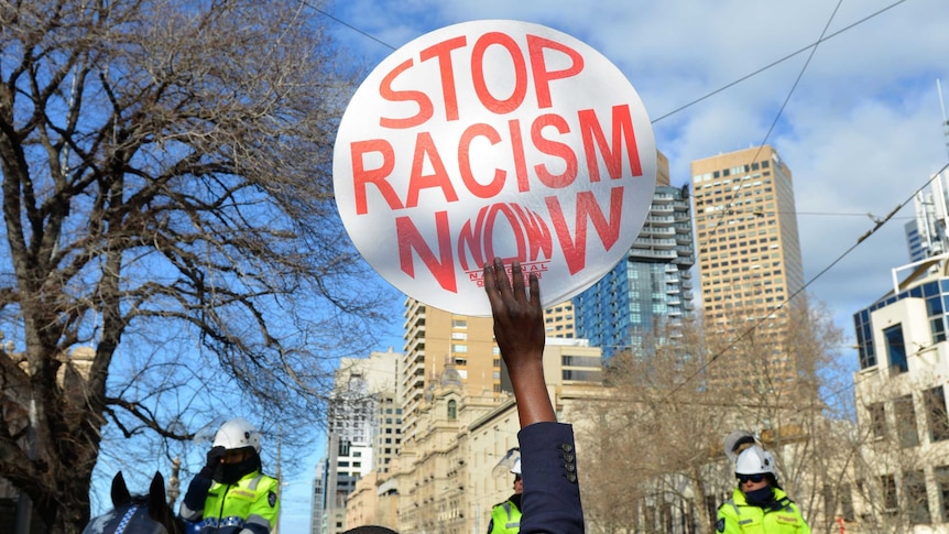 A man of colour holds up a sign saying "stop racism now" in front of Police on horses who are wearing hi viz
