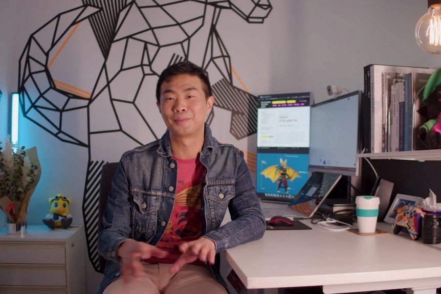 An Asian Australian man in his 30s sitting a white desk, pink tshirt and jean jacket, graphic image on the wall behind him