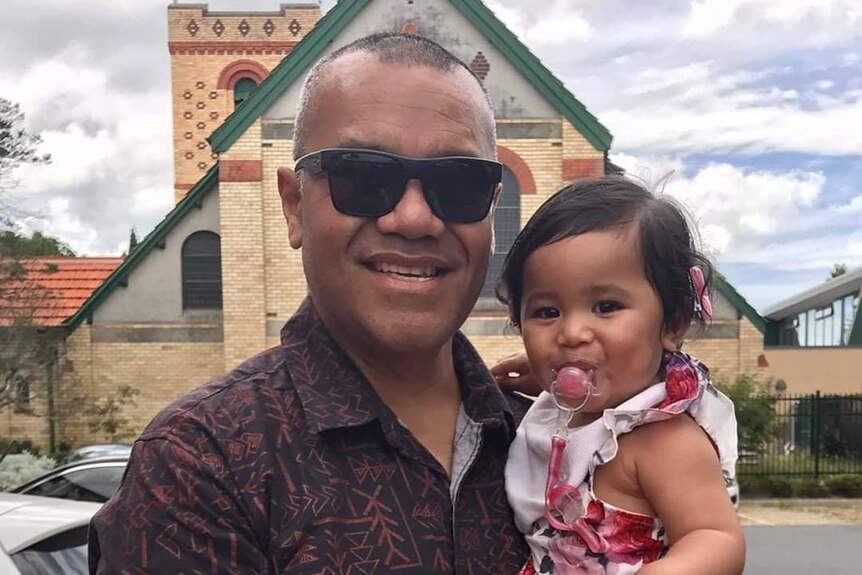 A man in sunglasses holds a little girl sucking on a dummy outside a church