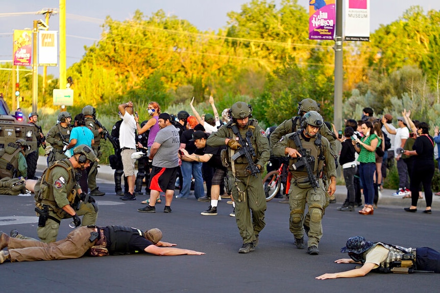 Heavily armed police wearing green stand over men face down on the ground wearing bulletproof vests.