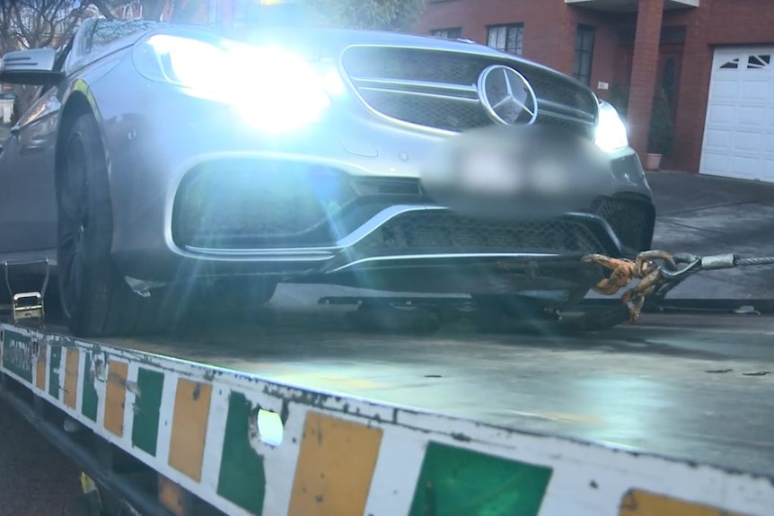 A silver Mercedes, with lights on, is pulled onto the tray of a tow truck.