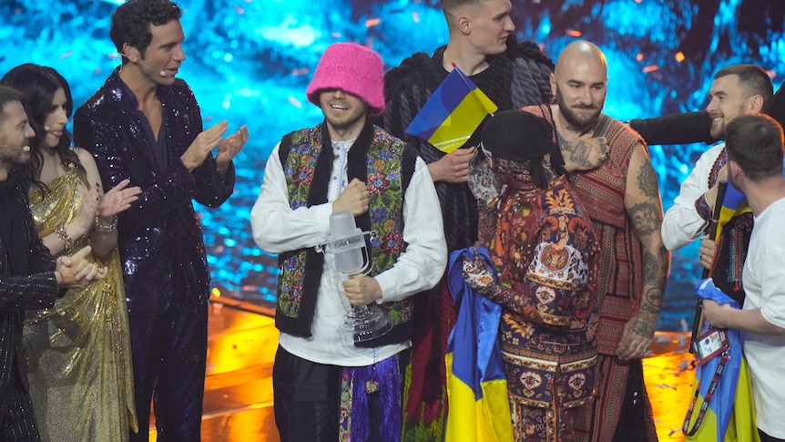 Kalush Orchestra holding Ukrainian flags, the fists to their hearts. One member holds the Eurovision trophy
