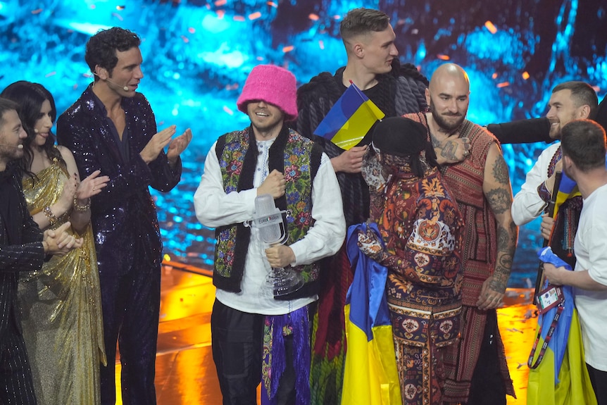 Kalush Orchestra holding Ukrainian flags, the fists to their hearts. One member holds the Eurovision trophy