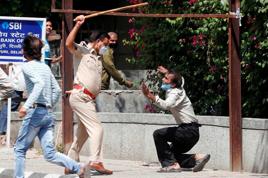 A police officer holds a cane above a crouching man in a face mask