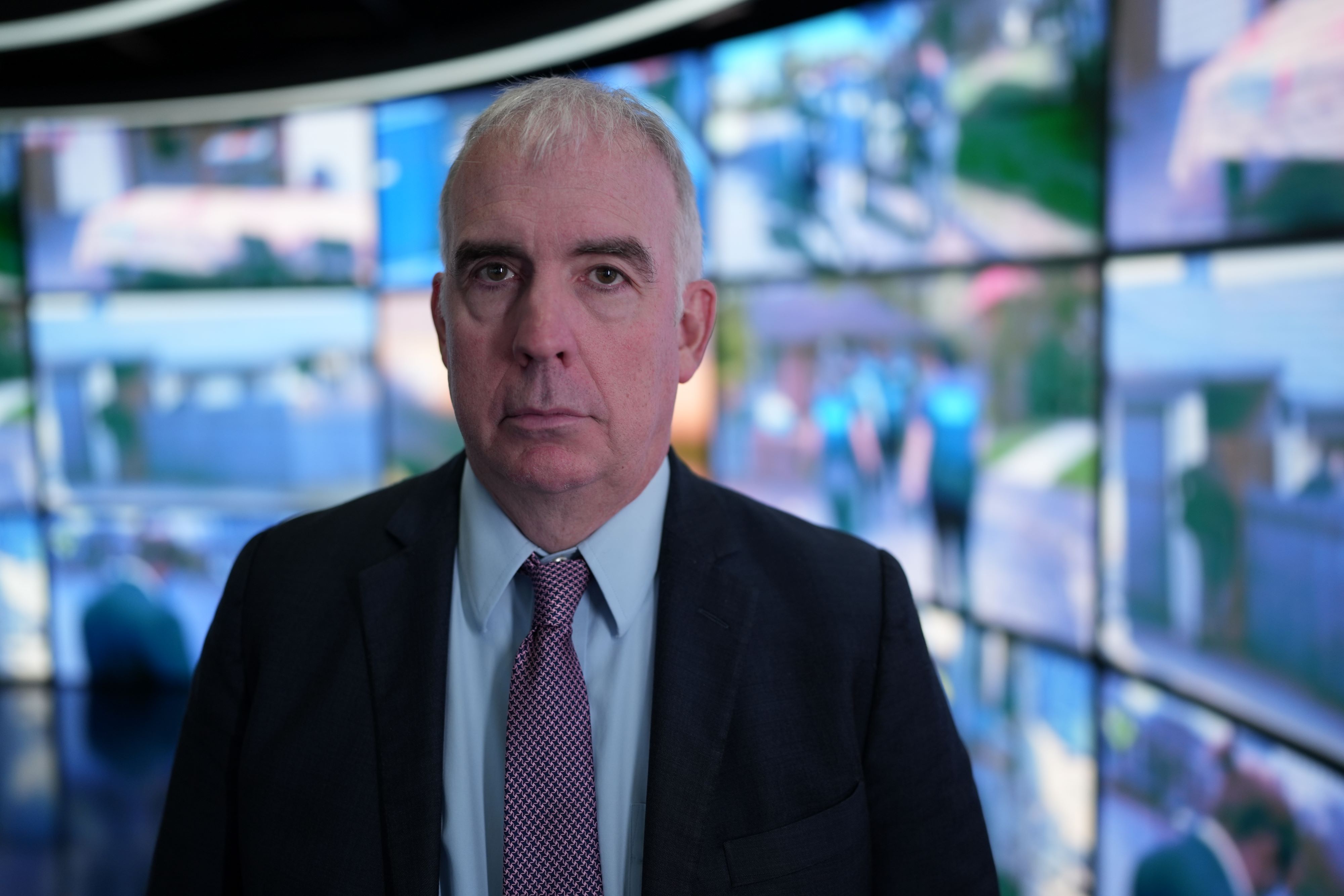 A man in a suit and tie looks into camera with a serious expression as he stands in front of a wall of video screens.