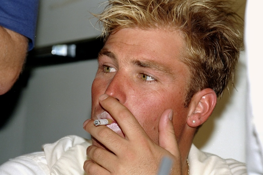 Shane Warne sits in a dressing room smoking a cigarette