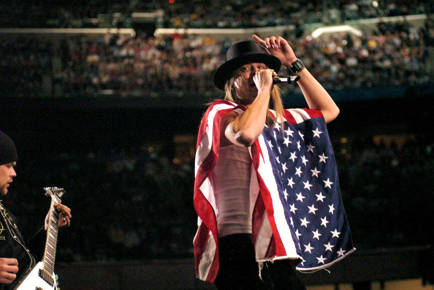 Kid Rock wearing the American flag as a poncho and holding a microphone