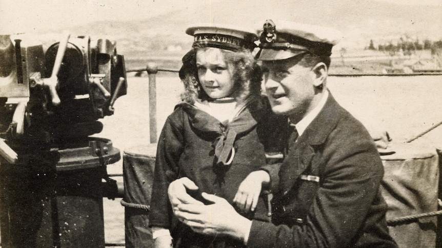 Girl on ship held by man crouched to her height. They both wear navy uniforms