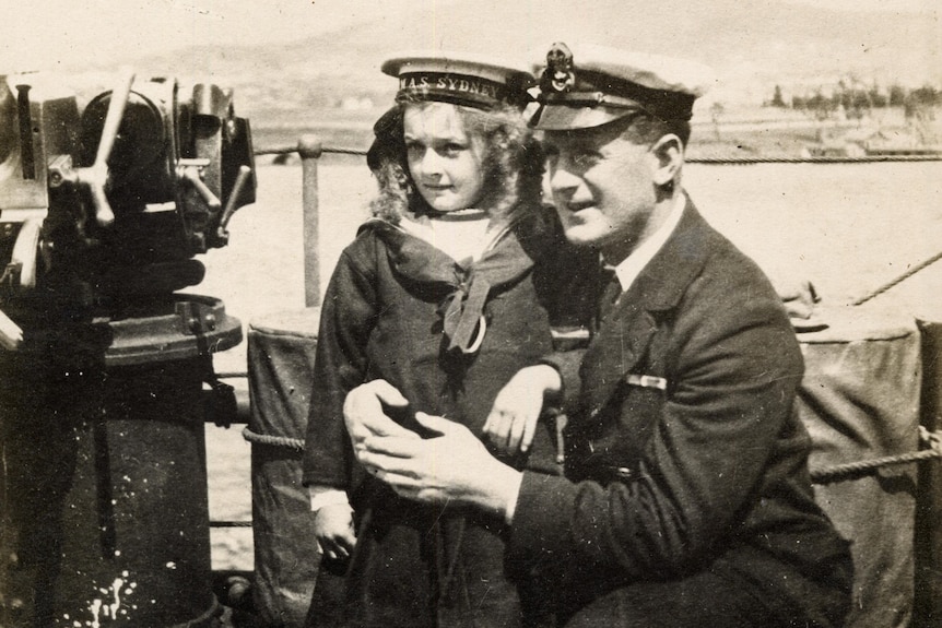 Girl on ship held by man crouched to her height. They both wear navy uniforms