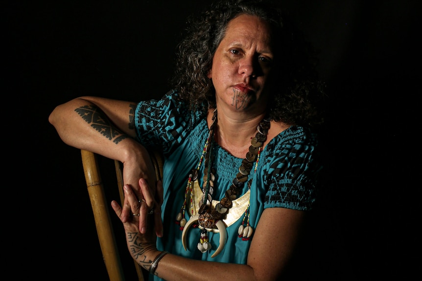 A woman with facial tattoos poses for a photograph against a dark background.