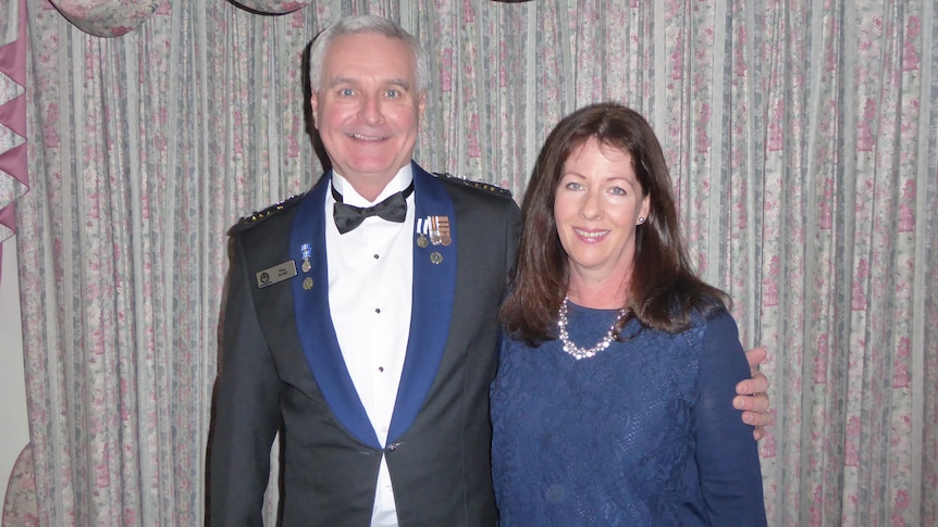 A man in a blue and black police suit with medals on its lapels with his arm around his wife, a woman with dark hair