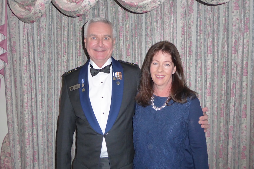 A man in a blue and black police suit with medals on its lapels with his arm around his wife, a woman with dark hair