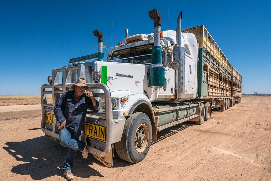 A man in an Akubra leans on a semi-trailer on a dusty red road.